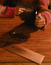 A person in a fuzzy pink and brown sweater holds a lighter over a dark incense holder on a wooden table, with a branded incense packet labeled 'MOCHA MAGIC INCENSE' in the foreground.