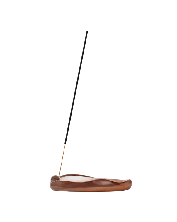 A single incense stick stands upright in a curved, polished brown wooden holder on a plain white background.