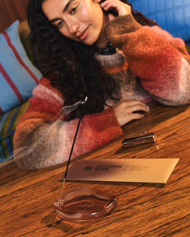 A woman in a fuzzy multicolor sweater leans over a wooden table, with DedCool Incense + Holder set in Mocha Magic burning in front of her