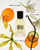 A clear glass perfume bottle with a black cap in the scent 'Orange Blossom'. The bottle is surrounded by artistic botanical elements including an orange blossom, white orchid, vanilla pod, and citrus slices on a clean white background.