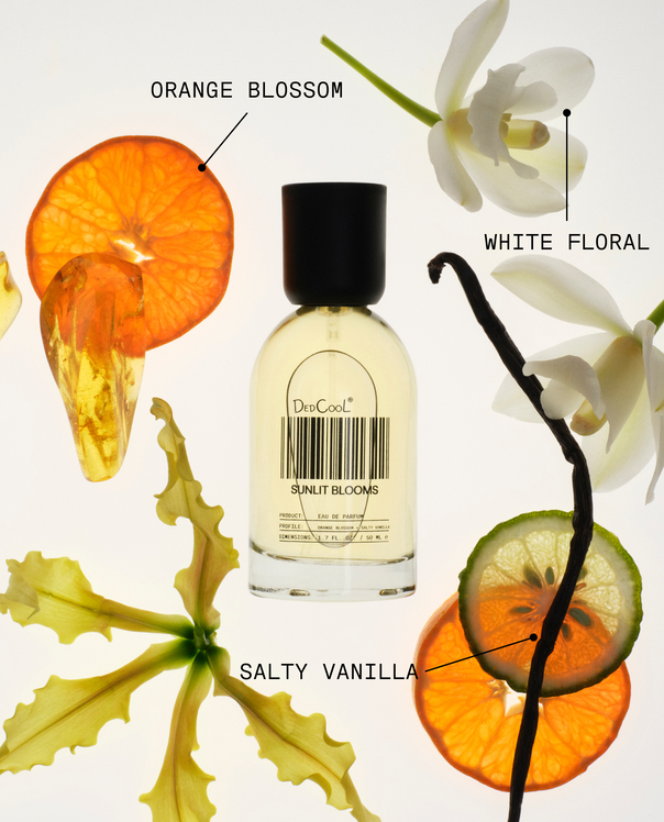 A clear glass perfume bottle with a black cap in the scent 'Orange Blossom'. The bottle is surrounded by artistic botanical elements including an orange blossom, white orchid, vanilla pod, and citrus slices on a clean white background.