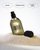A clear bottle of DedCool 'Sunlit Blooms' fragrance or body product filled with pale yellow liquid, shown standing in a pile of white granules with its black cap beside it.