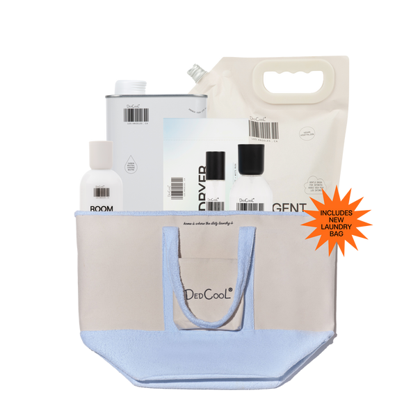 A DedCool travel toiletry bag set in light blue and beige, shown filled with travel-sized toiletries including a white bottle, a white tin, a white pouch, and a folded paper towel. An orange starburst sticker on the right reads 'INCLUDES NEWLY DESIGNED BAG'.