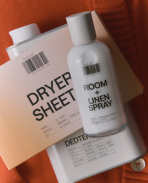 A white bottle of DedCool Room + Linen Spray placed on top of a beige dryer sheet and a white laundry bag, set against an orange textured fabric background.